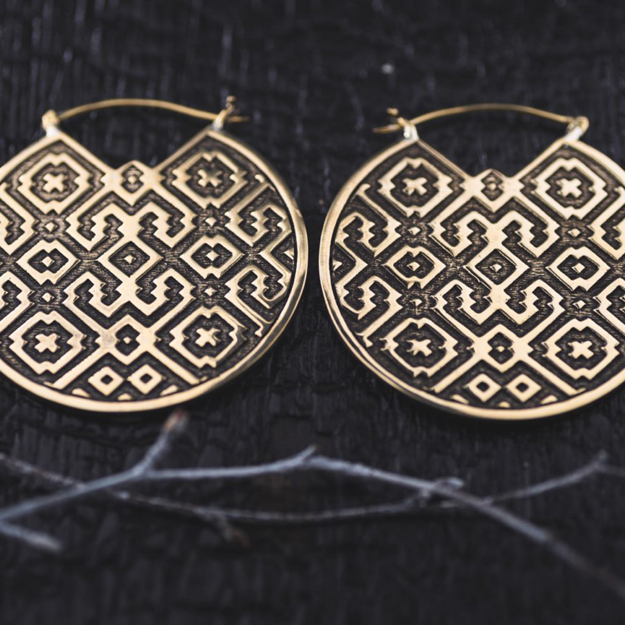 The AREVALO Shipibo Oversized Disc Hoop Earrings in Gold by Kaya Movement feature large, circular brass design etched with intricate Sacred geometry Shipibo patterns. These 14 gauge tribal earrings rest against a textured dark background, showcasing symmetrical shapes and dots that contribute to their bold, culturally-inspired look.